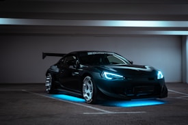 A sleek, modified sports car with aggressive styling sits in a dimly lit, empty parking garage. The car has bright blue LED lights highlighting its underside and headlights, giving it a futuristic appearance. It features a large rear wing and shiny alloy wheels, emphasizing its performance look.
