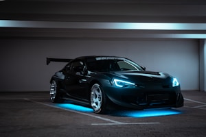 A sleek, modified sports car with aggressive styling sits in a dimly lit, empty parking garage. The car has bright blue LED lights highlighting its underside and headlights, giving it a futuristic appearance. It features a large rear wing and shiny alloy wheels, emphasizing its performance look.