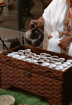 A person wearing traditional attire is pouring liquid from a dark, ornate pot into numerous small white cups arranged on a wooden chest. The chest has a textured pattern, and the scene is set on a green surface.