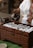 A person wearing traditional attire is pouring liquid from a dark, ornate pot into numerous small white cups arranged on a wooden chest. The chest has a textured pattern, and the scene is set on a green surface.
