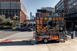 A vibrant food cart is stationed on an urban street corner, featuring a variety of bright food images on display. The cart offers hot dogs, chili, and other fast food items with colorful signage. Nearby, a street with cars and a bus is visible, with tall buildings framing the background under a clear blue sky.