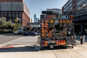A vibrant food cart is stationed on an urban street corner, featuring a variety of bright food images on display. The cart offers hot dogs, chili, and other fast food items with colorful signage. Nearby, a street with cars and a bus is visible, with tall buildings framing the background under a clear blue sky.