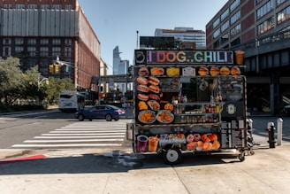 A vibrant food cart is stationed on an urban street corner, featuring a variety of bright food images on display. The cart offers hot dogs, chili, and other fast food items with colorful signage. Nearby, a street with cars and a bus is visible, with tall buildings framing the background under a clear blue sky.