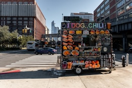 A vibrant food cart is stationed on an urban street corner, featuring a variety of bright food images on display. The cart offers hot dogs, chili, and other fast food items with colorful signage. Nearby, a street with cars and a bus is visible, with tall buildings framing the background under a clear blue sky.