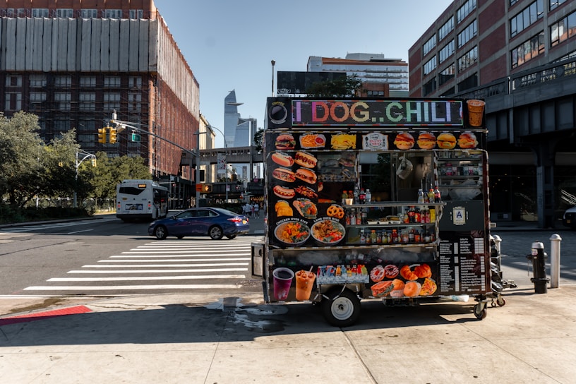 A vibrant food cart is stationed on an urban street corner, featuring a variety of bright food images on display. The cart offers hot dogs, chili, and other fast food items with colorful signage. Nearby, a street with cars and a bus is visible, with tall buildings framing the background under a clear blue sky.