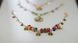 A colorful bracelet with circular beads in various hues such as red, orange, blue, and green arranged in a sequence. Gold-tone circular charms are interspersed between the beads, adding an elegant touch. The bracelet is laid out on a smooth, light surface, with a soft focus on similar accessories in the background.