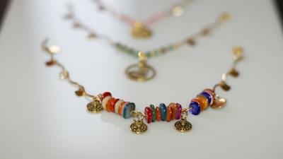 A colorful bracelet with circular beads in various hues such as red, orange, blue, and green arranged in a sequence. Gold-tone circular charms are interspersed between the beads, adding an elegant touch. The bracelet is laid out on a smooth, light surface, with a soft focus on similar accessories in the background.