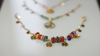 A colorful bracelet with circular beads in various hues such as red, orange, blue, and green arranged in a sequence. Gold-tone circular charms are interspersed between the beads, adding an elegant touch. The bracelet is laid out on a smooth, light surface, with a soft focus on similar accessories in the background.
