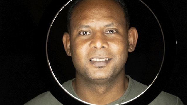 A person with a warm smile is illuminated within a circular frame against a dark background. The lighting emphasizes their facial features with a glowing effect.