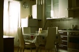 A quiet kitchen featuring modern white cabinetry, a refrigerator, and kitchen chairs around a table with a tablecloth. A large window with sheer white curtains allows natural light to fill the room. A wall clock hangs near the refrigerator, and household items are placed on the counters.