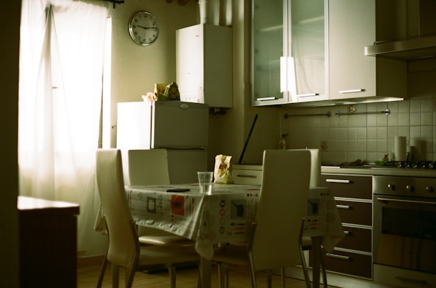 A quiet kitchen featuring modern white cabinetry, a refrigerator, and kitchen chairs around a table with a tablecloth. A large window with sheer white curtains allows natural light to fill the room. A wall clock hangs near the refrigerator, and household items are placed on the counters.
