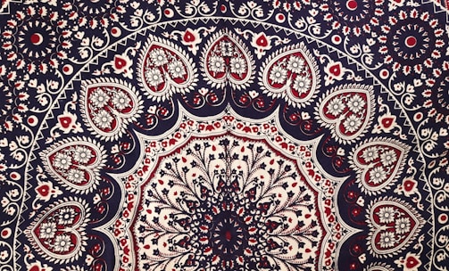 Intricate mandala pattern featuring a series of concentric circles adorned with floral and paisley motifs. The design includes heart-shaped elements filled with detailed floral patterns, primarily in shades of red, black, and white. The symmetry and repetition create a sense of balance and harmony.
