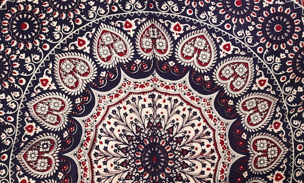 Intricate mandala pattern featuring a series of concentric circles adorned with floral and paisley motifs. The design includes heart-shaped elements filled with detailed floral patterns, primarily in shades of red, black, and white. The symmetry and repetition create a sense of balance and harmony.