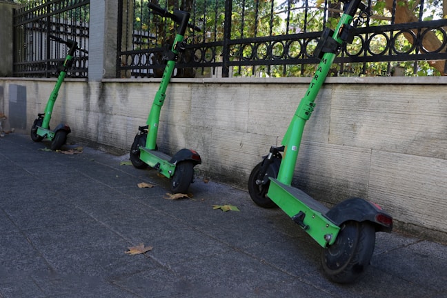 Three green electric scooters are lined up against a stone wall on a gray sidewalk. Fallen leaves are scattered around, and a decorative metal fence is visible in the background with green foliage beyond.