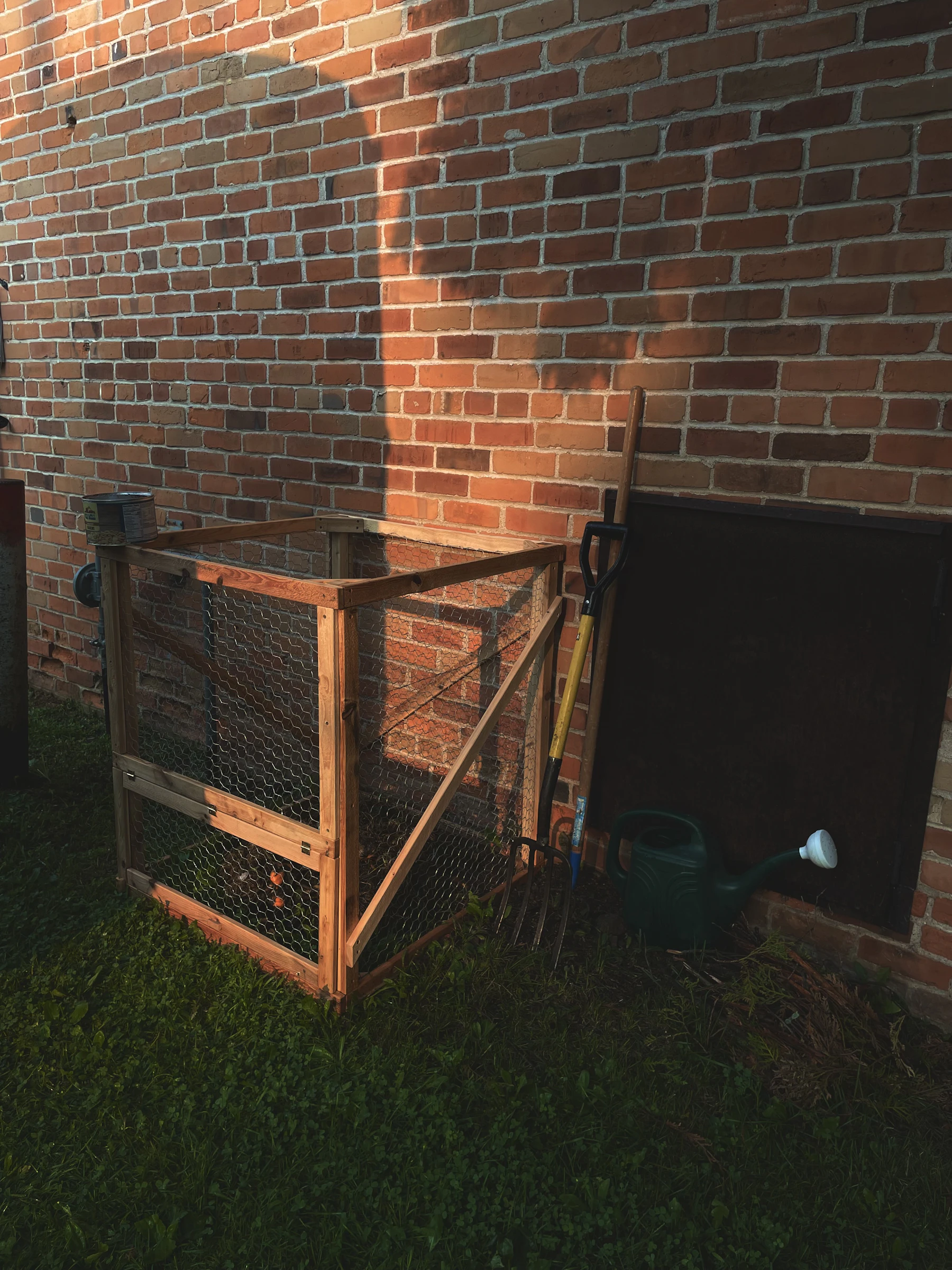 A square bin of wooden rails and wire mesh on a lawn beside a brick wall, with a garden fork and a green watering can leaning next to it