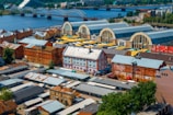 A vibrant cityscape with a mix of old and new architecture. The scene includes several large market halls with arched roofs, surrounded by industrial and residential buildings. A river flows in the background with a distinctive arched bridge crossing it. The area is bustling, with a variety of colors from the roofs and awnings.