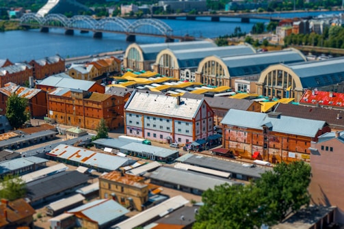 A vibrant cityscape with a mix of old and new architecture. The scene includes several large market halls with arched roofs, surrounded by industrial and residential buildings. A river flows in the background with a distinctive arched bridge crossing it. The area is bustling, with a variety of colors from the roofs and awnings.