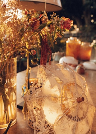 A delicate lace parasol rests gently on a table, surrounded by a warm and cozy ambiance. Soft sunlight filters through, highlighting a bouquet of dried flowers and a few glowing candles that add a touch of tranquility and charm.