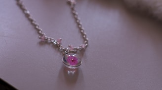A delicate necklace with a silver chain featuring a pendant containing a pink flower encased in glass. The chain has small pink beads evenly spaced along its length, creating a subtle, feminine aesthetic.