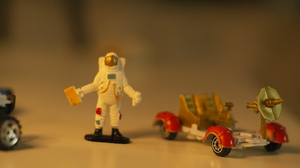 A small figurine of an astronaut in a white spacesuit with colorful details stands beside a miniature lunar rover. The rover has red wheel covers and a dish antenna. The setting is softly lit, creating a warm and nostalgic ambiance.