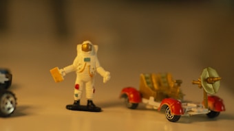 A small figurine of an astronaut in a white spacesuit with colorful details stands beside a miniature lunar rover. The rover has red wheel covers and a dish antenna. The setting is softly lit, creating a warm and nostalgic ambiance.