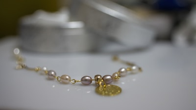 A delicate bracelet featuring pearls and gold links is placed on a smooth surface. The focus is on the jewelry, with cylindrical shapes and a metal pendant creating a luxurious feel. The background is softly blurred, highlighting the elegance of the piece.