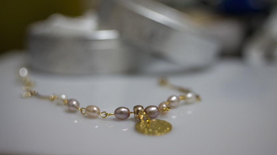 A delicate bracelet featuring pearls and gold links is placed on a smooth surface. The focus is on the jewelry, with cylindrical shapes and a metal pendant creating a luxurious feel. The background is softly blurred, highlighting the elegance of the piece.