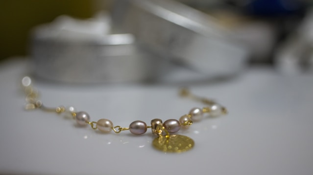 A delicate bracelet featuring pearls and gold links is placed on a smooth surface. The focus is on the jewelry, with cylindrical shapes and a metal pendant creating a luxurious feel. The background is softly blurred, highlighting the elegance of the piece.