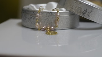An elegant, decorative bracelet with pearls and a gold charm is draped over an ornate silver box, which has a floral pattern. The box lid is slightly ajar, revealing a soft, white interior.