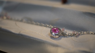 A close-up of a delicate silver chain bracelet featuring a round pink gemstone set within a clear bezel. The bracelet appears to be laid on a soft, light-colored fabric with subtle patterns.