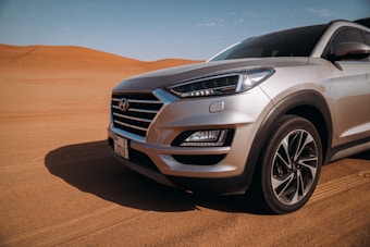 A silver SUV is parked on a sandy terrain with undulating sand dunes in the background under a clear blue sky.