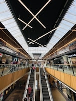 A bustling shopping mall with an escalator at the center leading to the upper floor. Shoppers are seen ascending and descending, creating a lively atmosphere. The ceiling features modern, geometric lighting fixtures, and the stores on either side have clean, modern storefront designs.