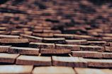 Roughly cut wooden shingles are laid out in a repeating pattern, creating an intricate and textured surface. The shingles, with visible natural grain and cracks, are weathered, suggesting they have been exposed to the elements for some time. The focus is on the shingles in the foreground, while the background becomes progressively blurred.