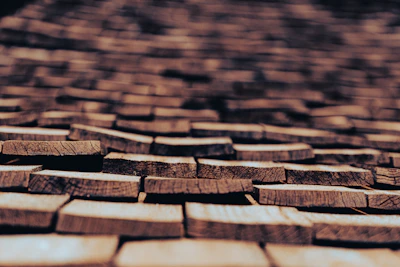 Roughly cut wooden shingles are laid out in a repeating pattern, creating an intricate and textured surface. The shingles, with visible natural grain and cracks, are weathered, suggesting they have been exposed to the elements for some time. The focus is on the shingles in the foreground, while the background becomes progressively blurred.