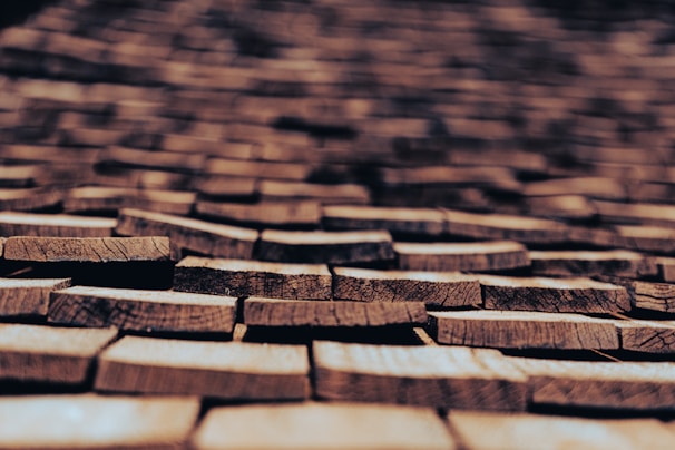 Roughly cut wooden shingles are laid out in a repeating pattern, creating an intricate and textured surface. The shingles, with visible natural grain and cracks, are weathered, suggesting they have been exposed to the elements for some time. The focus is on the shingles in the foreground, while the background becomes progressively blurred.