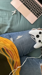 A close-up view of a person's lap with a pair of ripped blue jeans and a colorful striped shirt. On the lap rests a white gaming controller. A rose gold laptop is open to the left, set on a blue-green surface. Nearby, a pair of white wireless earbuds is lying on the surface, and a white earbud cable is partially visible.