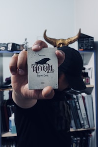A person holds a deck of cards labeled 'Ravn Playing Cards' with a crow image on the front. The individual is partially obscured, wearing dark clothing and a cap, and stands in a room filled with books and shelves in the background. There is a decorative horned object positioned behind their head.
