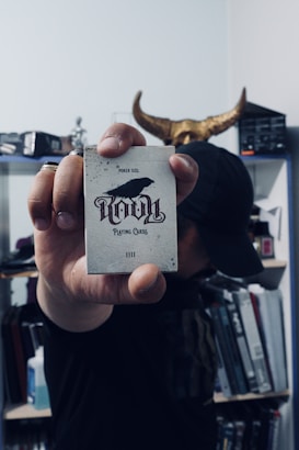 A person holds a deck of cards labeled 'Ravn Playing Cards' with a crow image on the front. The individual is partially obscured, wearing dark clothing and a cap, and stands in a room filled with books and shelves in the background. There is a decorative horned object positioned behind their head.