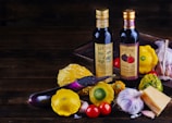 A rustic arrangement featuring two bottles of Lucini oil placed on a wooden surface surrounded by various fresh ingredients. There are colorful squashes and a purple eggplant, as well as garlic bulbs, cherry tomatoes, and a chunk of cheese, creating a vibrant and earthy display.