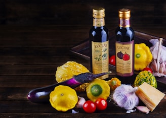A rustic arrangement featuring two bottles of Lucini oil placed on a wooden surface surrounded by various fresh ingredients. There are colorful squashes and a purple eggplant, as well as garlic bulbs, cherry tomatoes, and a chunk of cheese, creating a vibrant and earthy display.