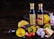 A rustic arrangement featuring two bottles of Lucini oil placed on a wooden surface surrounded by various fresh ingredients. There are colorful squashes and a purple eggplant, as well as garlic bulbs, cherry tomatoes, and a chunk of cheese, creating a vibrant and earthy display.