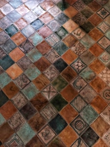 The image displays a pattern of colorful ceramic tiles arranged in a grid. The tiles feature various intricate designs and symbols, including floral, geometric, and abstract motifs. The tiles appear to be artistically handcrafted, with a mix of glossy and matte finishes.