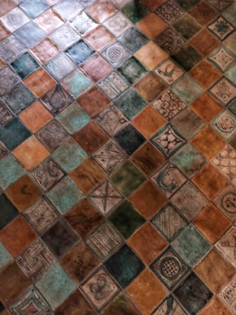 The image displays a pattern of colorful ceramic tiles arranged in a grid. The tiles feature various intricate designs and symbols, including floral, geometric, and abstract motifs. The tiles appear to be artistically handcrafted, with a mix of glossy and matte finishes.