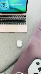 A laptop with a rose gold finish displays a music streaming app on its screen. An AirPods case is placed on a light gray fabric surface next to purple fabric material. A gaming controller partially visible at the bottom right corner.