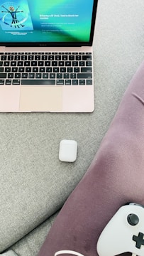 A laptop with a rose gold finish displays a music streaming app on its screen. An AirPods case is placed on a light gray fabric surface next to purple fabric material. A gaming controller partially visible at the bottom right corner.