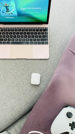 A laptop with a rose gold finish displays a music streaming app on its screen. An AirPods case is placed on a light gray fabric surface next to purple fabric material. A gaming controller partially visible at the bottom right corner.