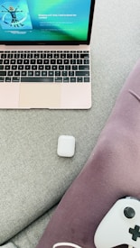 A laptop with a rose gold finish displays a music streaming app on its screen. An AirPods case is placed on a light gray fabric surface next to purple fabric material. A gaming controller partially visible at the bottom right corner.