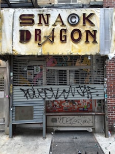 A storefront with a yellow and brown sign that reads 'Snack Dragon.' The storefront has a metal roll-up gate with graffiti on it, and the window behind the gate is covered with various colorful stickers and posters. The building appears to be old and worn, with visible cracks in the walls and peeling paint. The surrounding area is urban, with a concrete sidewalk and a brick wall to the right.