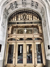An ornate entrance of a historical building features a grand arched doorway with intricate carvings and decorative elements. The polished brass doors are reflective, showcasing a person taking a photo and the street behind them. Above the doors, the text 'United States Realty Building' is prominently displayed on an ornate arch.