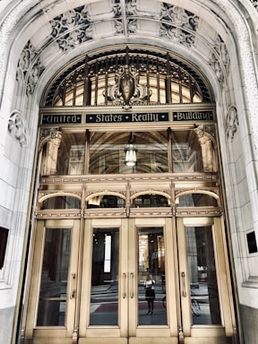 An ornate entrance of a historical building features a grand arched doorway with intricate carvings and decorative elements. The polished brass doors are reflective, showcasing a person taking a photo and the street behind them. Above the doors, the text 'United States Realty Building' is prominently displayed on an ornate arch.